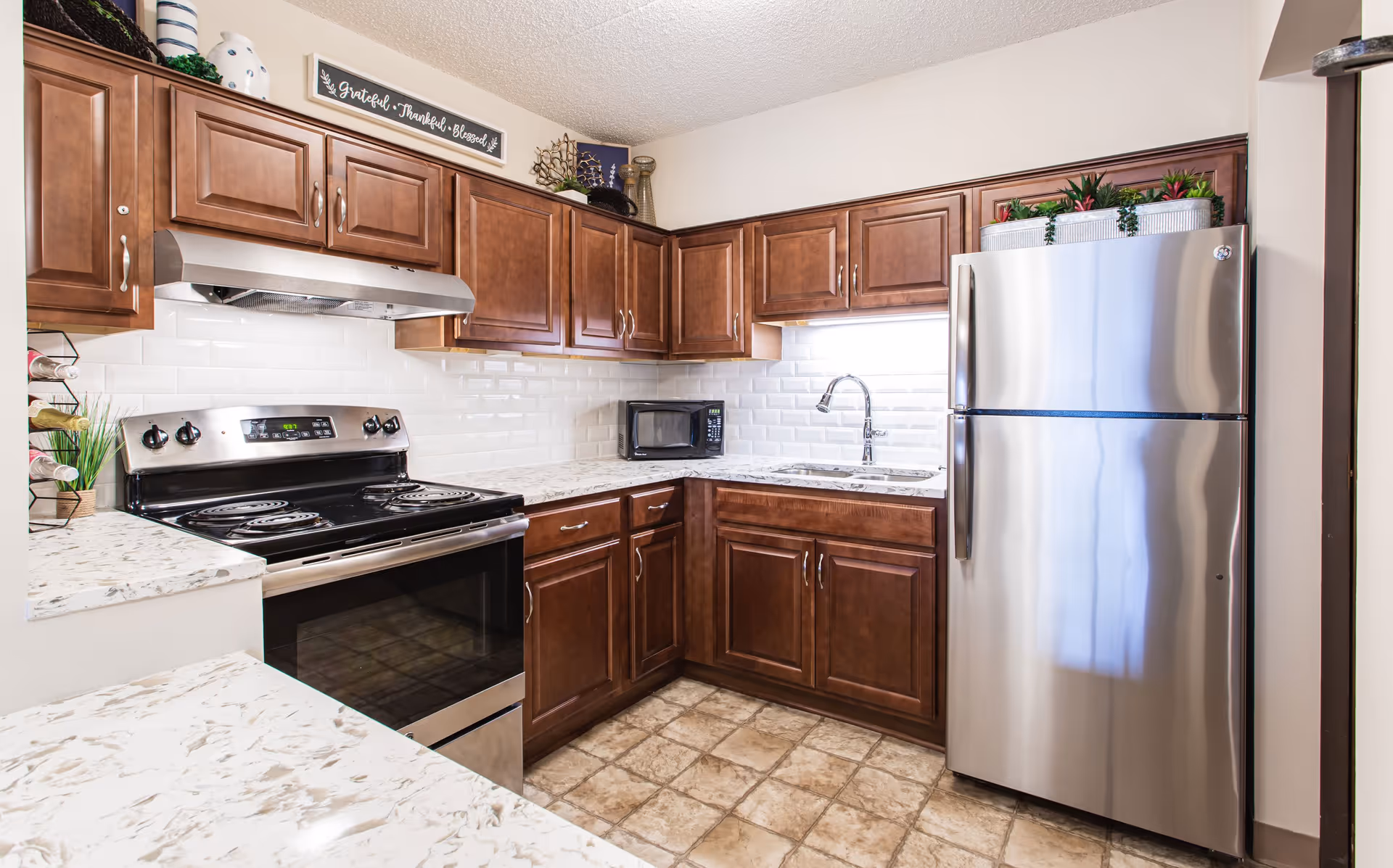 A modern kitchen with brown wooden cabinets, a stainless steel electric stove with oven, a stainless steel refrigerator, a microwave on the countertop, a sink with a faucet, and decorative plants on top of the refrigerator. The backsplash is white subway tile and the floor has beige tiles.