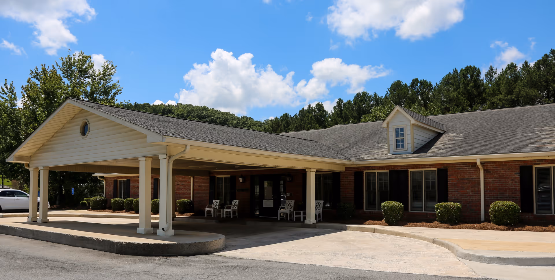 Front entrance of a single-story brick senior living facility with a covered porte-cochère and chairs under the canopy against a blue sky.