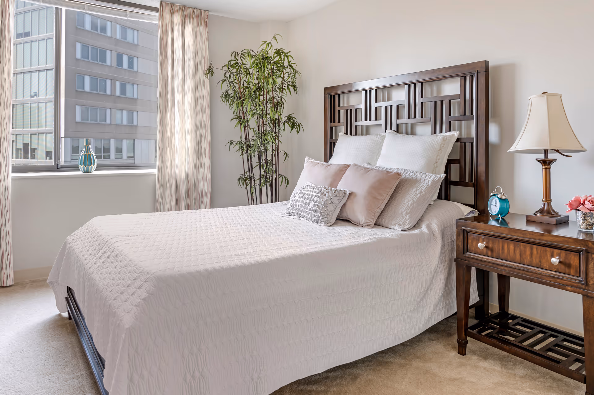 A neatly made bed with a white quilted bedspread and multiple pillows in a bedroom. The bed has a dark wooden headboard with a geometric design. Next to the bed is a wooden nightstand with a lamp, a blue alarm clock, and a small vase with pink flowers. A tall green plant is placed in the corner near a window with light-colored curtains, showing a view of a building outside.