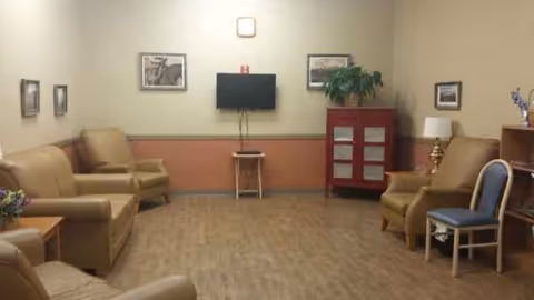 Senior living facility lounge with tan seating, a wall TV, plant, and framed pictures.