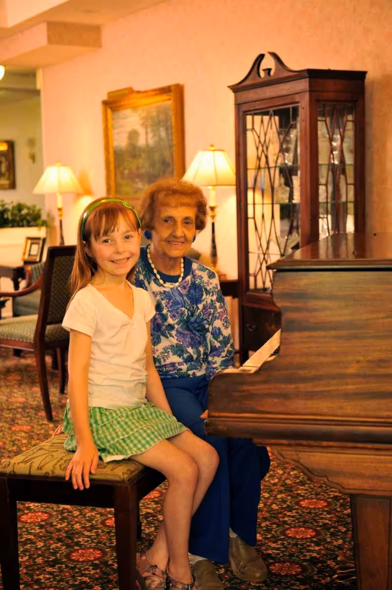 An elderly woman and a young girl sitting together on a piano bench in a warmly lit living room. The elderly woman is wearing a blue floral blouse and blue pants, while the girl is dressed in a white shirt and green checkered skirt. Behind them, there is a wooden display cabinet with glass doors, two table lamps, and framed artwork on the wall.