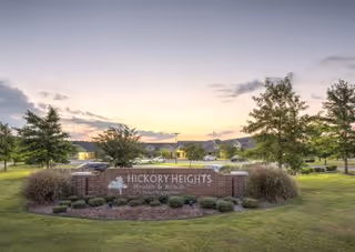 Brick entrance sign reading "Hickory Heights Health & Rehab" set in a landscaped lawn with trees and the facility buildings in the background at sunset.