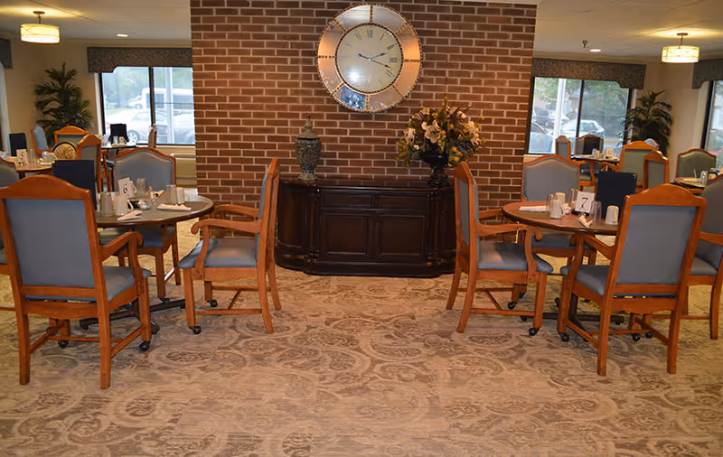 Dining area in an assisted living facility with wooden tables and chairs upholstered in blue fabric. The room features a patterned carpet, a brick accent wall with a large decorative clock, a dark wooden sideboard with a vase and a flower arrangement, and windows with valances letting in natural light.