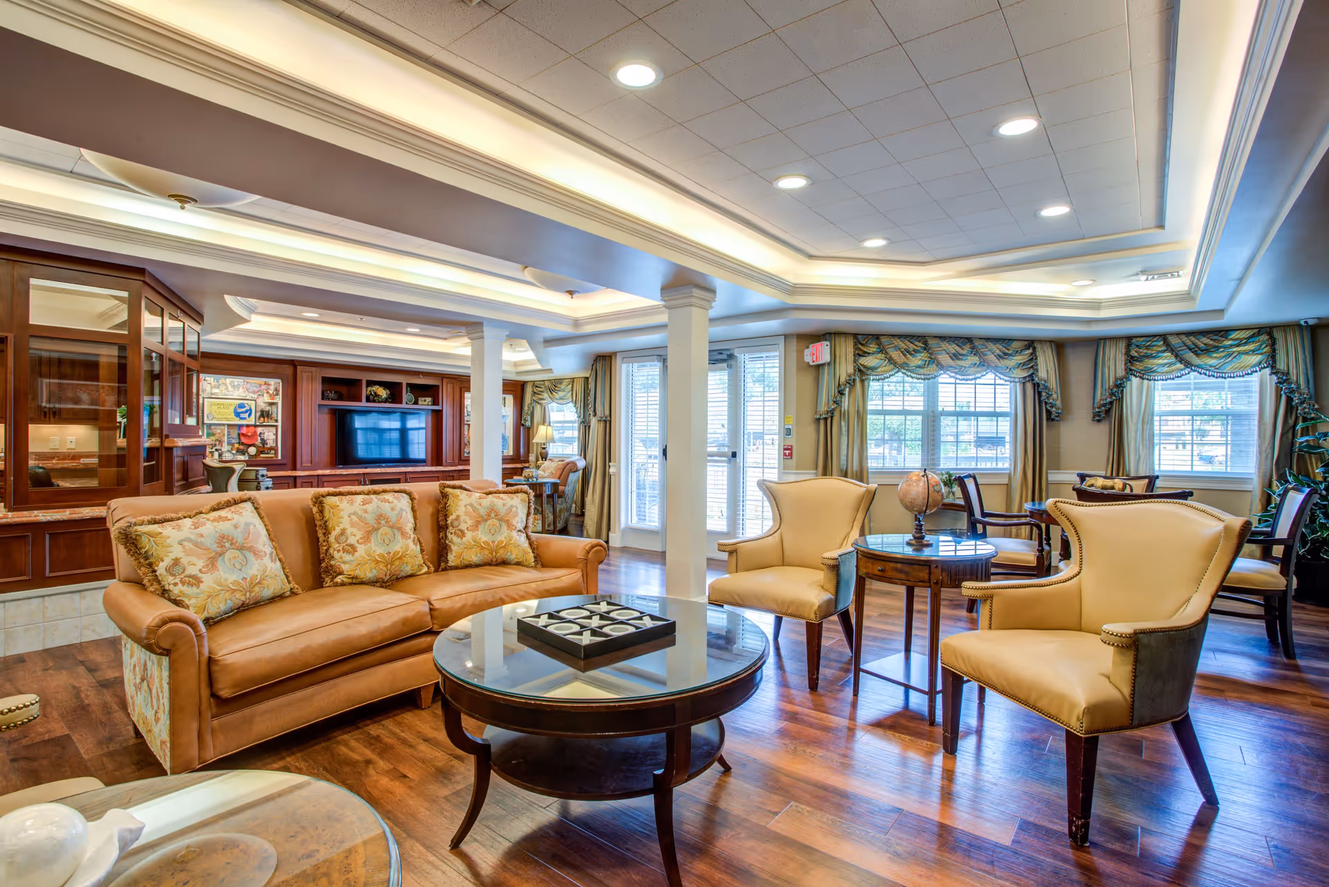 A bright, upscale communal lounge with a leather sofa, upholstered armchairs, coffee tables, built-in TV cabinet, and large draped windows.