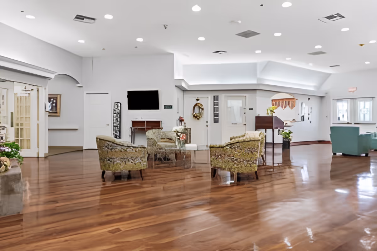 A spacious and well-lit common area with polished wooden floors, several upholstered armchairs arranged around a glass coffee table, and a flat-screen TV mounted on the wall. The room has white walls, recessed ceiling lights, and decorative elements including plants and a wall mirror. There are multiple doors and windows visible, contributing to an open and inviting atmosphere.