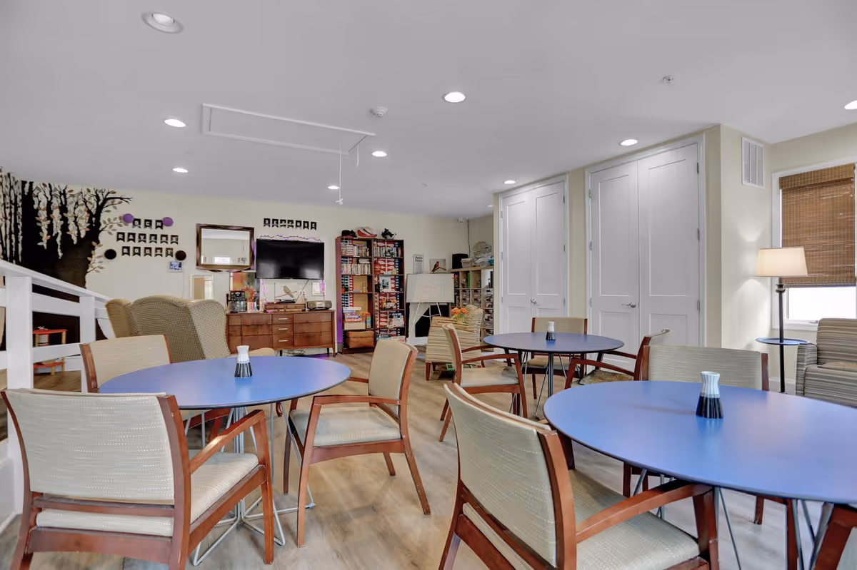 A common area in Broad Creek Manor Assisted Living featuring several round tables with chairs arranged around them. The room has light-colored walls, wood flooring, and recessed ceiling lights. There is a TV mounted on the wall above a wooden cabinet, bookshelves filled with games and books, a whiteboard, and a floor lamp near a window with a bamboo shade. A decorative tree mural is painted on one wall.