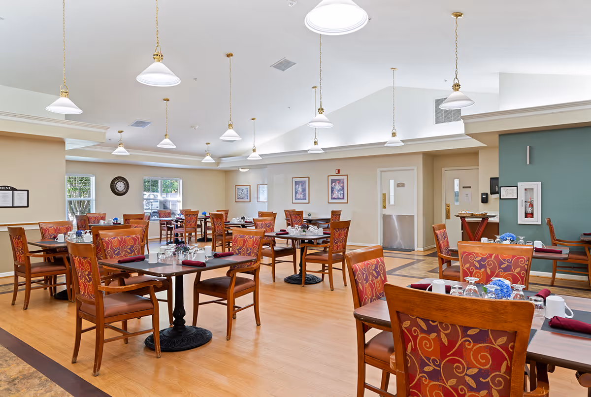 Spacious senior living dining room with multiple set tables and patterned chairs under pendant lights.