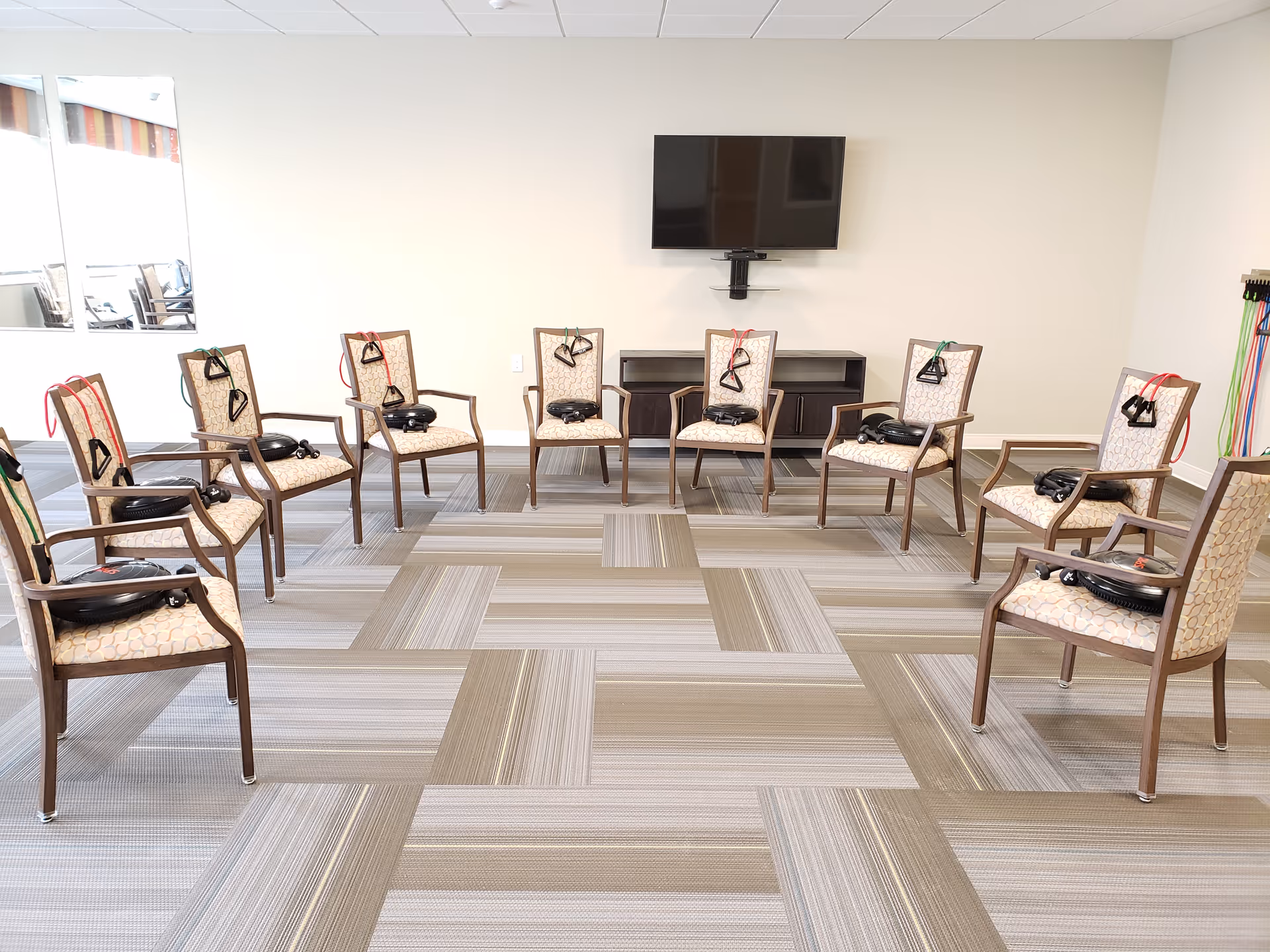 A bright room with ten chairs arranged in a circle, each chair equipped with resistance bands and exercise equipment. A flat-screen TV is mounted on the wall above a dark wooden cabinet. The floor has a patterned carpet with shades of beige and gray.