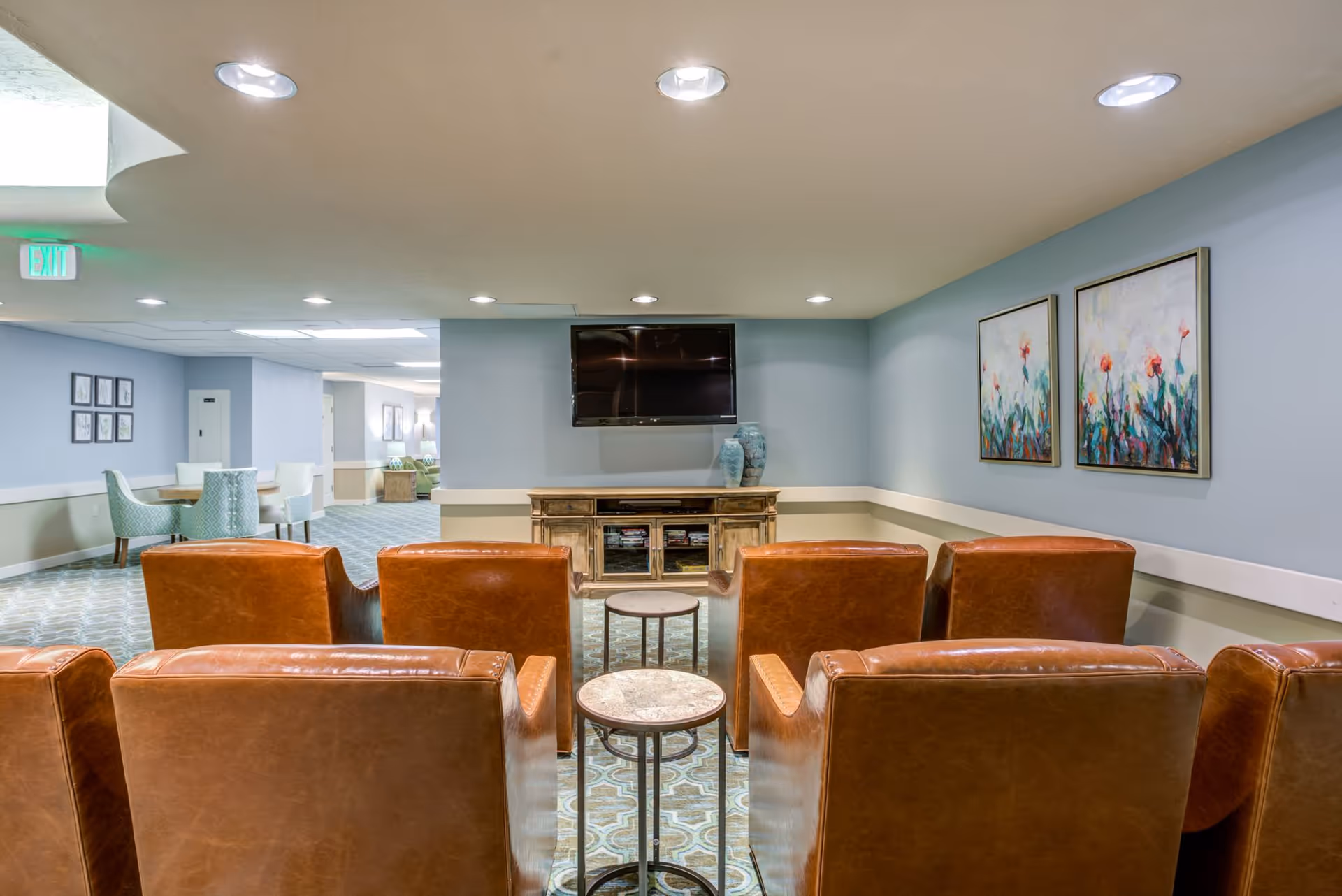 A cozy communal lounge with rows of brown leather chairs facing a wall-mounted TV and console, floral artwork on the wall, and small round side tables.