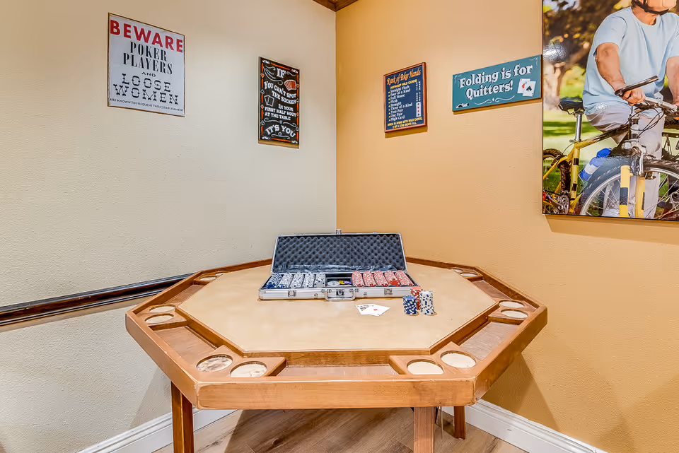 Octagonal poker table with poker chips and playing cards on top, set against walls decorated with poker-themed signs and a large photo of a person riding a bicycle.