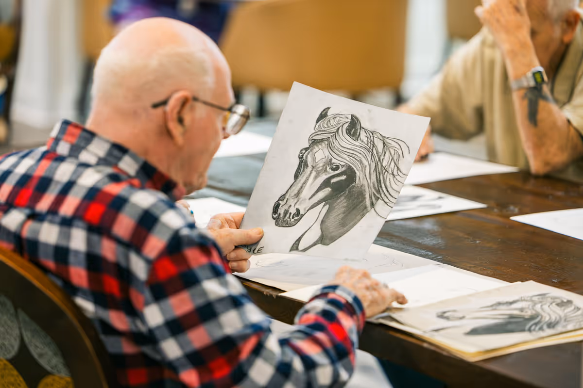 An elderly man wearing glasses and a red and black checkered shirt is sitting at a table, holding and looking at a detailed pencil drawing of a horse's head. Another elderly person is partially visible in the background, also seated at the table with drawings spread out in front of them.