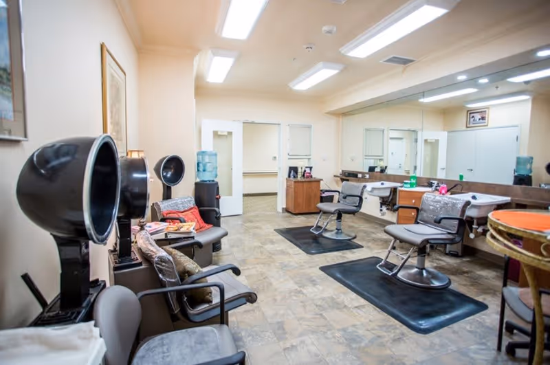 Interior view of a salon area in a senior living facility with hair drying chairs on the left, two salon chairs in front of a large mirror on the right, and a water cooler in the background. The room has tiled flooring and bright overhead lighting.