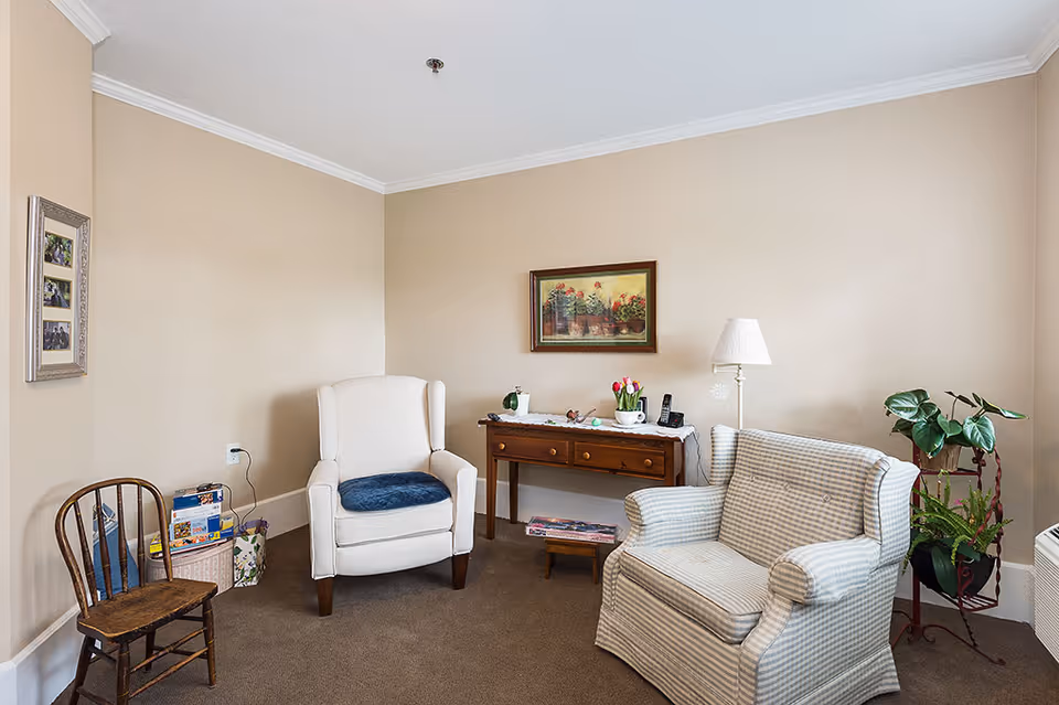A cozy sitting area in an assisted living facility featuring a white cushioned armchair with a blue cushion, a checkered upholstered armchair, a wooden chair, a wooden console table with decorative items including a lamp, flowers, and a phone, a framed painting on the beige wall, and a plant on a stand near the window.
