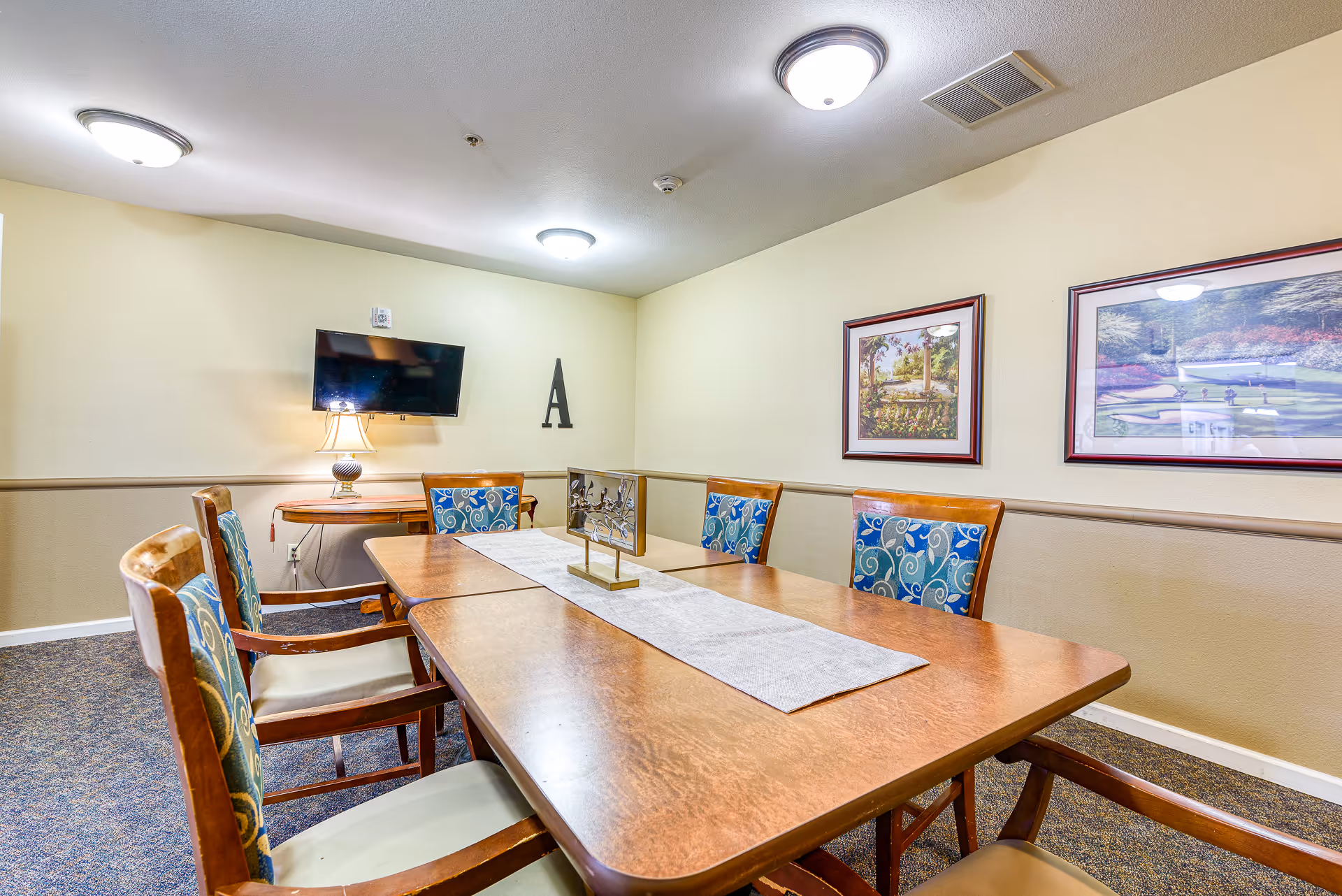 A small meeting or dining room with a rectangular wooden table surrounded by six wooden chairs with blue patterned cushions. The walls are painted beige with a chair rail molding. There are two framed pictures hanging on the wall and a flat-screen TV mounted on another wall above a small wooden table with a lamp. The room is lit by ceiling lights.