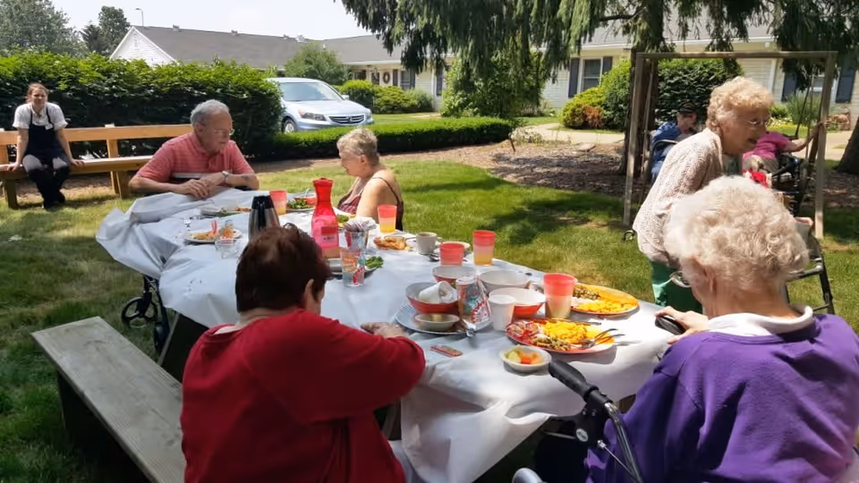 Several elderly residents sit around a picnic table eating outdoors on the lawn of a retirement village.