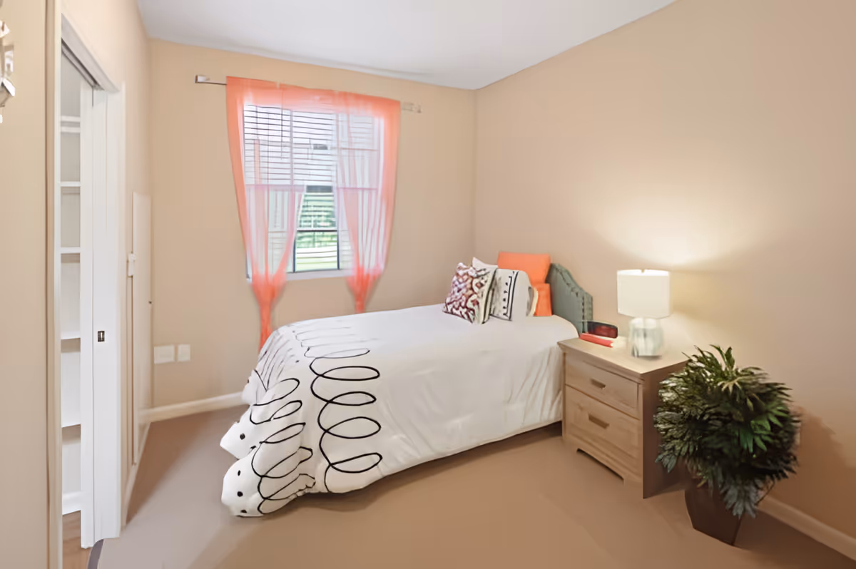 A small bedroom with beige walls and carpet. There is a single bed with a white bedspread featuring black swirl patterns, and several decorative pillows in orange and patterned designs. Next to the bed is a wooden nightstand with a lamp and a small red item. A green potted plant is placed on the floor beside the nightstand. A window with sheer orange curtains allows natural light into the room. A closet with sliding doors is visible on the left side of the image.