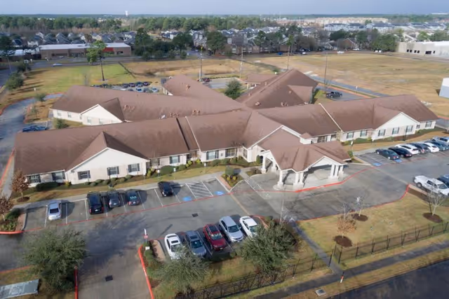 Aerial view of a single-story healthcare and rehabilitation center building with a brown roof and beige exterior walls. The building is surrounded by parking lots with several cars parked, some trees, and open grassy areas. The entrance features a covered drop-off area with columns. In the background, there are residential houses and other buildings.