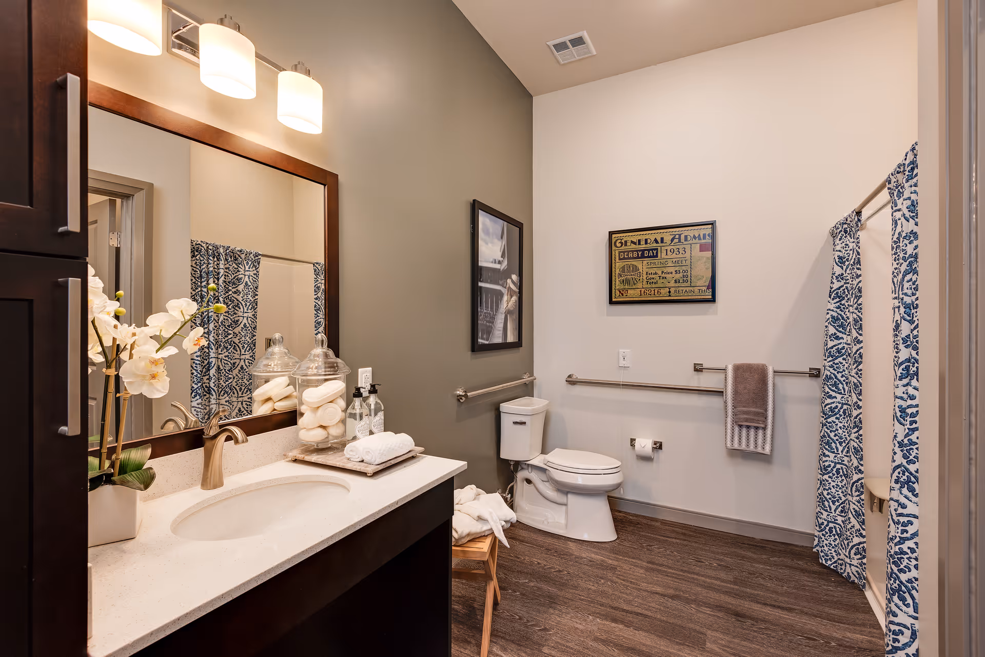 A clean and modern bathroom featuring a white countertop with a sink, a large mirror with three lights above it, a toilet with grab bars on the walls, a shower with a blue and white patterned curtain, and decorative items including a potted orchid and jars with soap and towels.