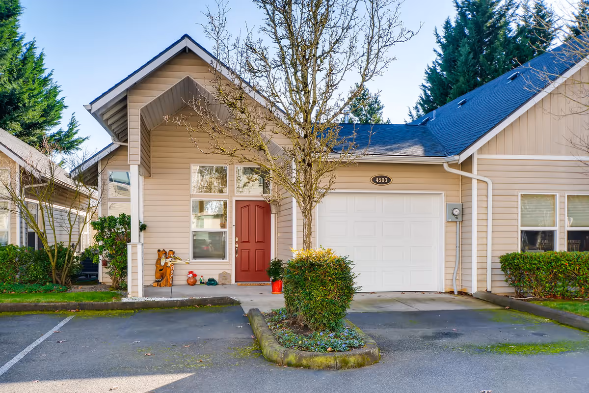 Exterior view of a beige single-story residential building with a red front door, a white garage door, and a small tree and shrubbery in front. The building has a dark blue roof and is surrounded by greenery and a paved driveway.
