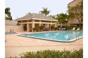 Outdoor swimming pool area with a small pool house or cabana, surrounded by palm trees and lounge chairs, under a clear sky.