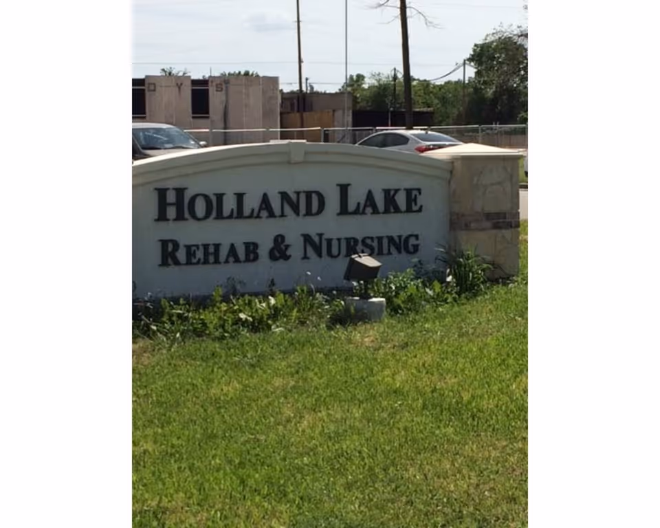 Outdoor stone sign for Holland Lake Rehab & Nursing surrounded by grass and some plants, with cars and a building in the background.