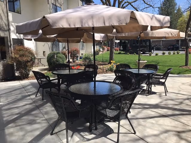 Outdoor patio area with round glass-top tables and black wicker chairs under large beige umbrellas, adjacent to a building with windows and surrounded by green grass and trees.