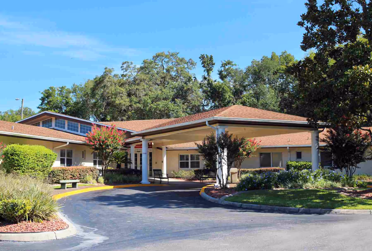 Exterior view of The Lodge Health & Rehabilitation Center showing a single-story building with a covered entrance, surrounded by trees, shrubs, and a clear blue sky.