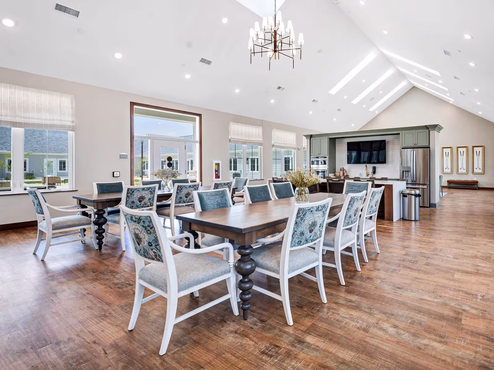 Bright and spacious dining room with wooden floors, multiple tables surrounded by white chairs with blue patterned cushions, large windows letting in natural light, and a kitchen area with green cabinetry, stainless steel appliances, and a mounted flat-screen TV.