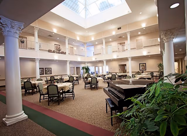 Spacious dining area with multiple tables covered with white tablecloths and green cushioned chairs arranged neatly. The room features tall white columns with ornate capitals, a large skylight ceiling allowing natural light to fill the space, and a black grand piano near some green plants in the foreground. The area has a carpeted floor and a second-floor balcony overlooking the dining space.