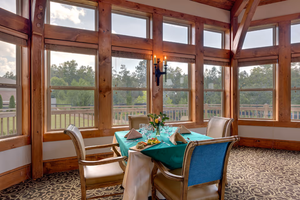 Sunlit dining area with a table set for four by large wood-framed windows overlooking greenery.