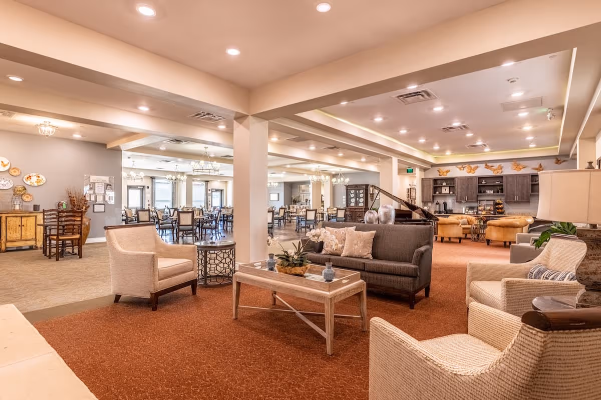 A spacious and well-lit common area in an assisted living facility featuring comfortable seating with armchairs and a sofa around a coffee table. In the background, there is a dining area with multiple tables and chairs, decorative wall plates, and a sideboard. The ceiling has recessed lighting and chandeliers, and the overall decor is warm and inviting.