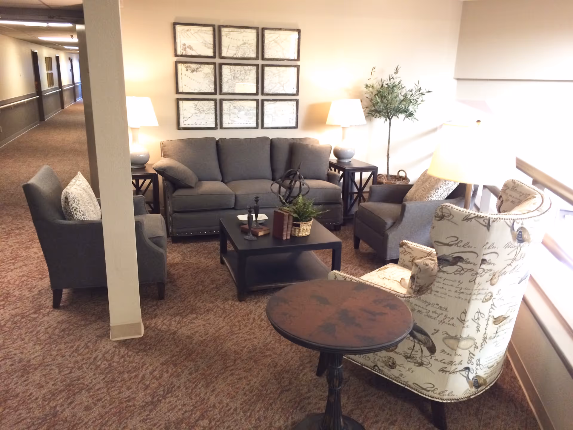 A cozy seating area in a senior living facility hallway featuring a gray sofa, two gray armchairs, and a patterned armchair with script and bird designs. The seating surrounds a black coffee table with decorative items, and there are two side tables with lamps. A potted plant is placed in the corner, and framed artwork hangs on the wall above the sofa. The area is carpeted and well-lit with natural and artificial light.