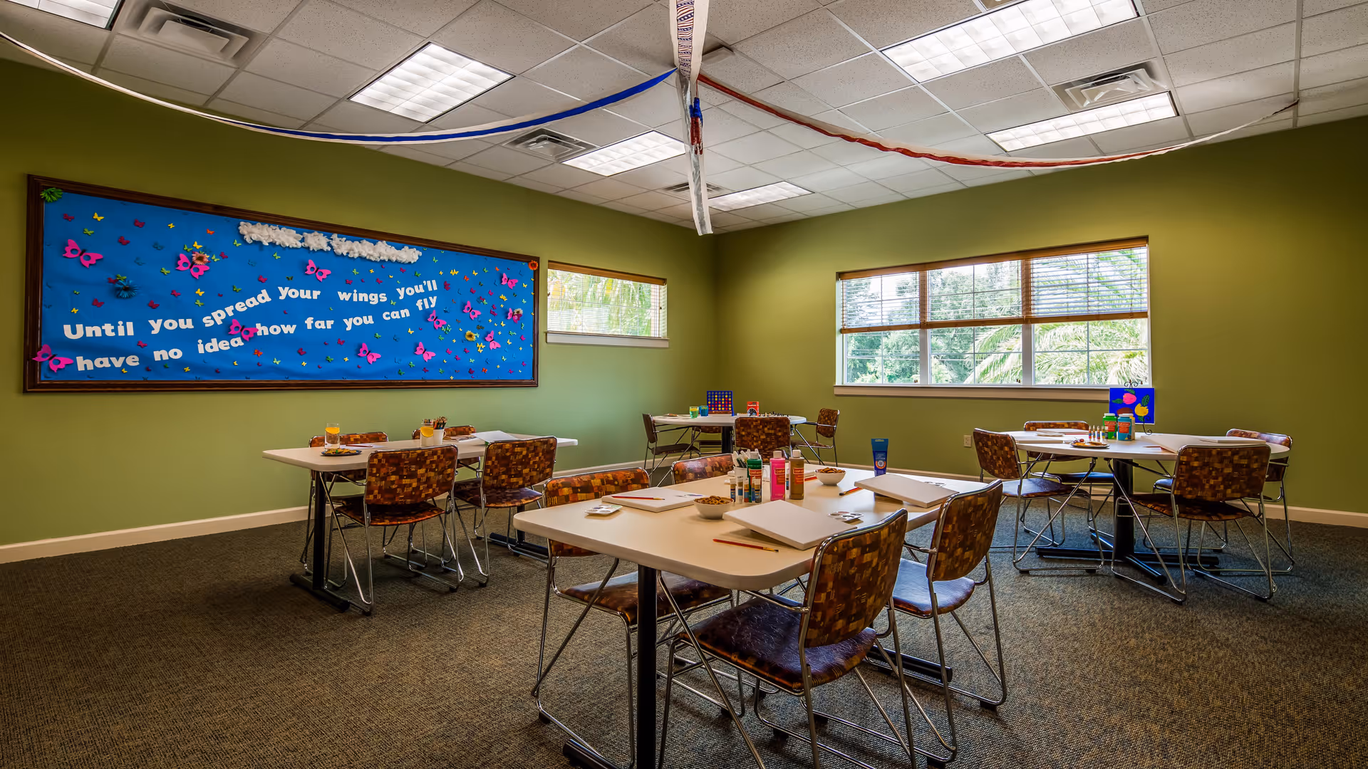 A well-lit activity room with green walls and carpeted floor featuring several tables and chairs arranged for group activities. The tables have art supplies like paint bottles, brushes, and paper. A large bulletin board on one wall displays a motivational quote with colorful butterfly decorations. Two windows let in natural light.