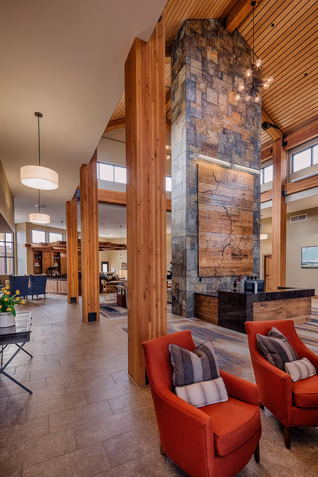 Spacious and well-lit common area in a senior living facility featuring tall wooden pillars, a large stone fireplace with a wooden map decoration, red armchairs with cushions, and various seating areas with tables and chairs. The ceiling is high with wooden paneling and modern light fixtures.