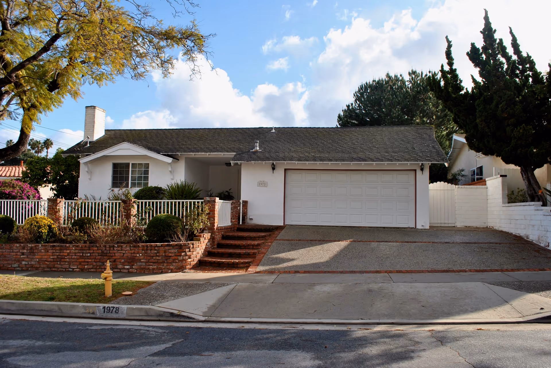 Front exterior view of a single-story house with a white garage door, brick steps leading to the entrance, a small garden with bushes and flowers, and a tree on the left side. The house number 1978 is visible on the curb and near the entrance.