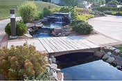 A landscaped outdoor area featuring a small wooden bridge over a clear water stream with rocks and a cascading waterfall surrounded by green shrubs and plants, with a paved walkway and lawn in the background.