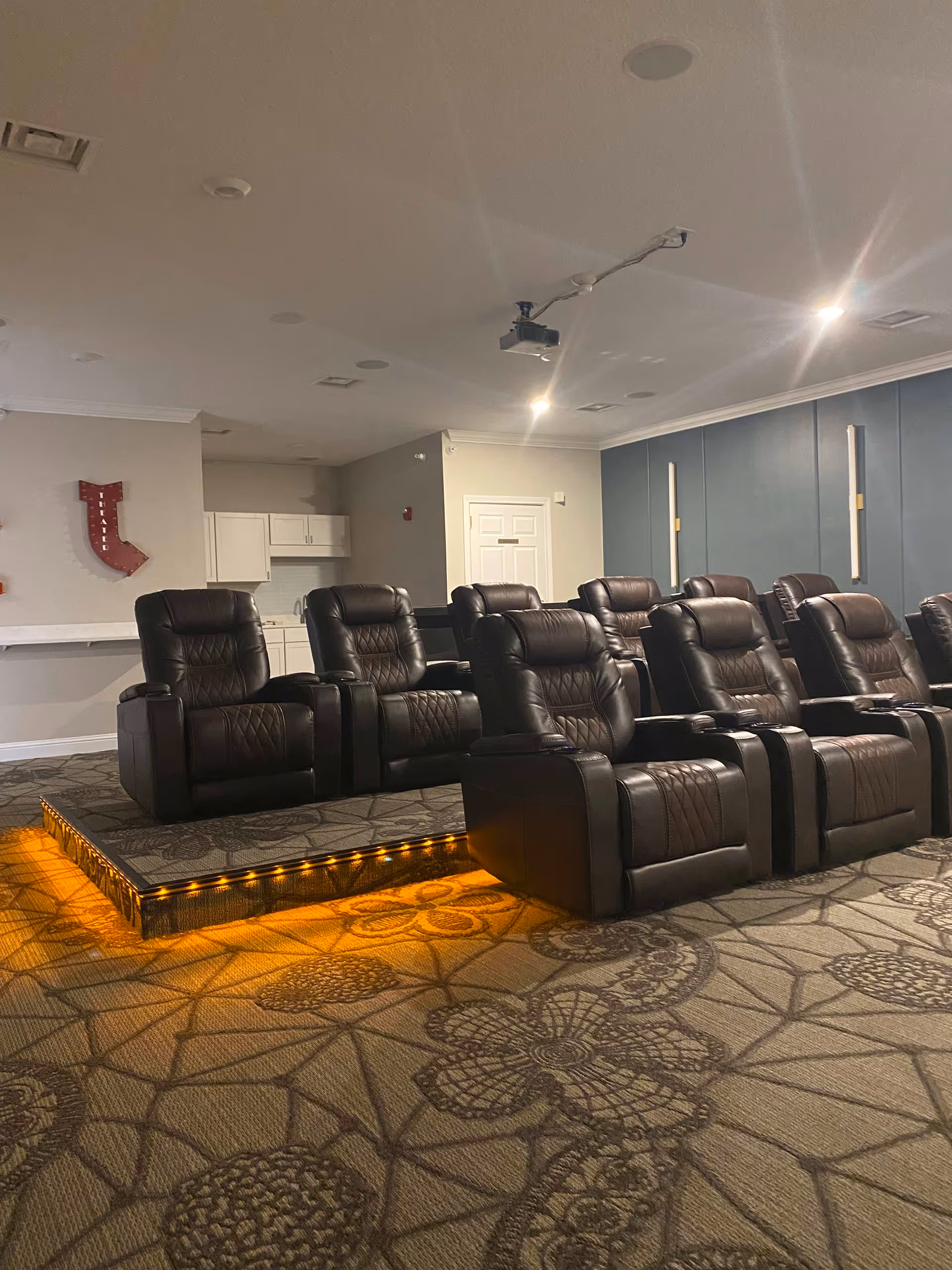 A small theater room with two rows of black leather recliner chairs on a carpeted floor with floral patterns. The front row is on the floor level, and the back row is elevated on a platform with orange lighting underneath. The walls are painted blue and beige, and there is a projector mounted on the ceiling. A kitchenette with white cabinets and a sink is visible in the background.