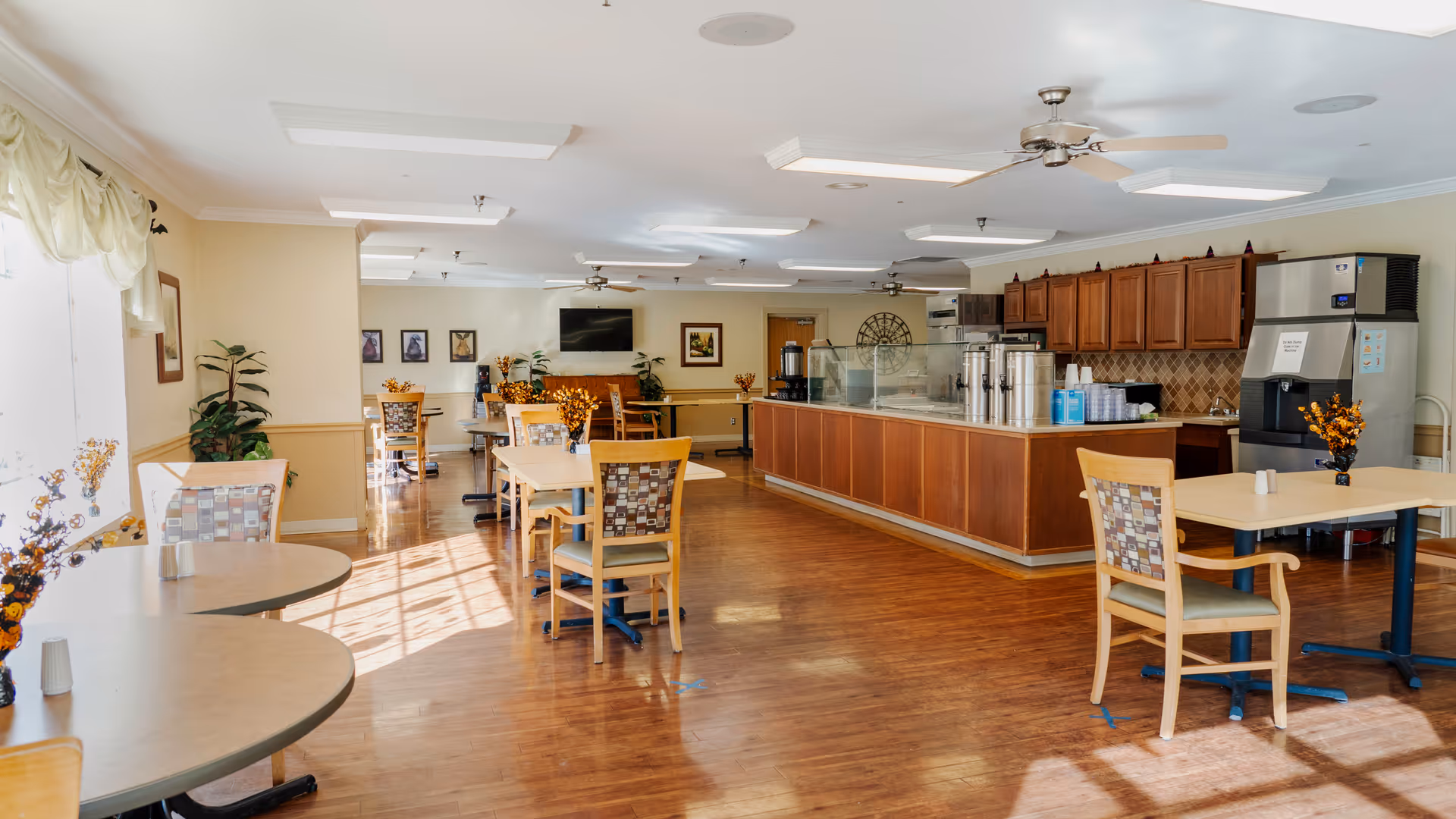 A bright dining area in a senior living facility with several tables and chairs arranged neatly. The room features wooden flooring, a serving counter with coffee dispensers, a large ice machine, and a television mounted on the far wall. The space is decorated with plants and framed pictures, and sunlight streams in through large windows with light curtains.