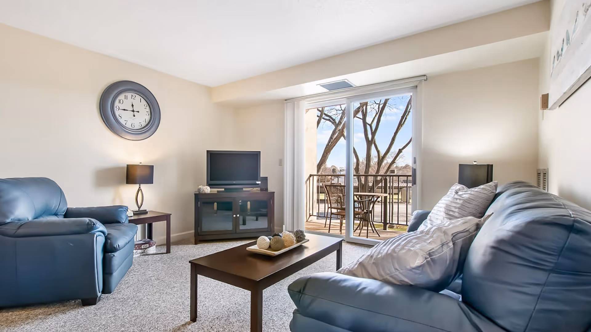 A cozy living room with a blue leather sofa and armchair, a wooden coffee table with decorative balls, a TV on a dark wooden stand, two table lamps, a wall clock, and a sliding glass door leading to a balcony with outdoor seating and trees visible outside.