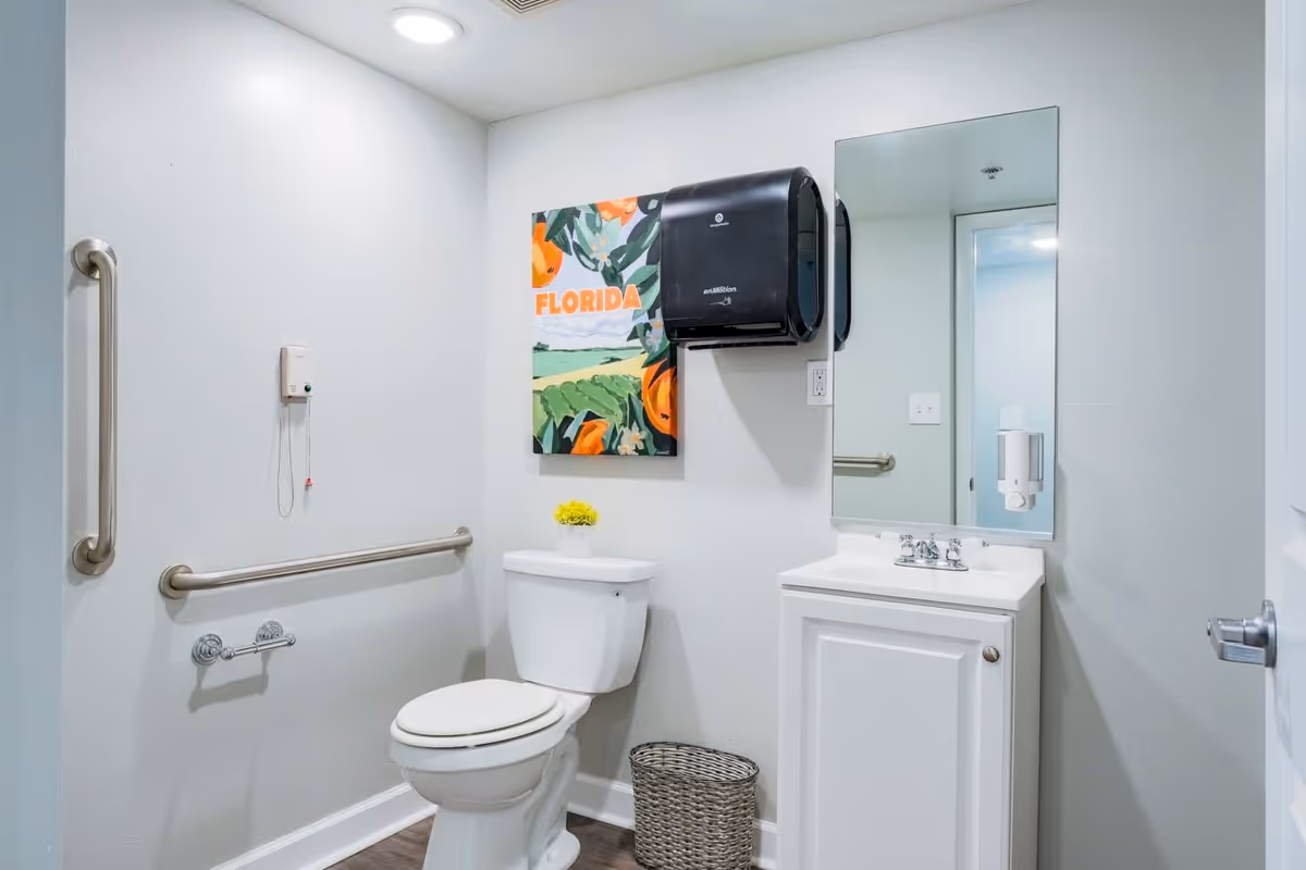 A clean and accessible bathroom with a white toilet, a white sink with a mirror above it, stainless steel grab bars on the walls, a black paper towel dispenser, a small waste basket, and a colorful painting on the wall that says 'FLORIDA'.