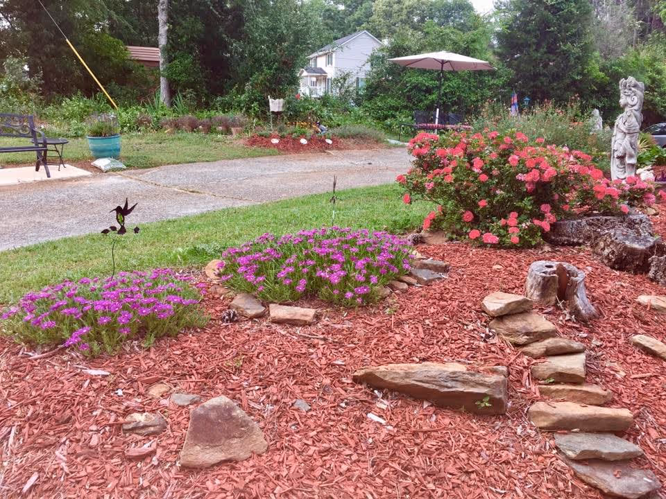 A landscaped outdoor garden area with red mulch, stone decorations, and flower beds containing purple and pink flowers. There is a paved driveway in the background, a bench on the left side, a patio umbrella with seating, and a white house partially visible behind trees and bushes.
