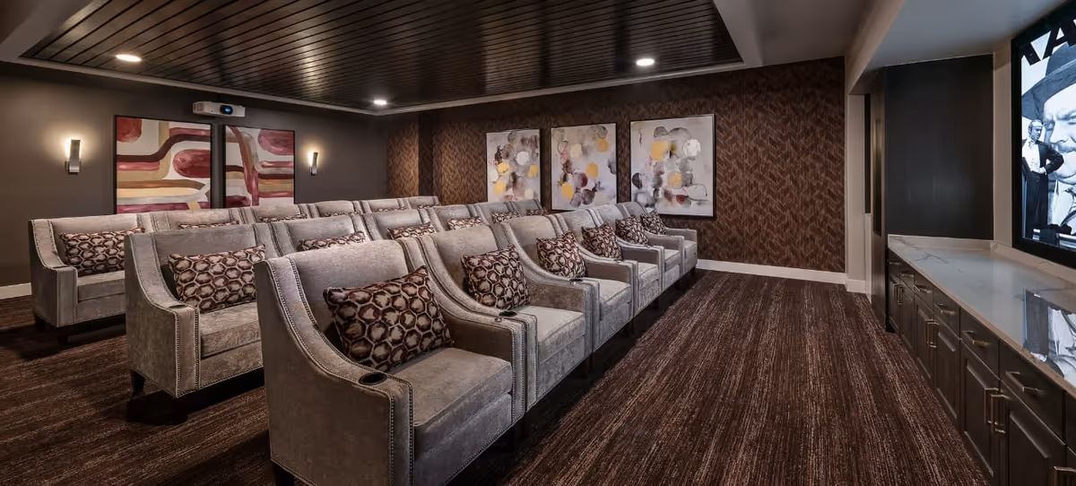 A cozy home theater room with two rows of plush gray armchairs, each with patterned throw pillows. The room features dark wood paneling on the ceiling, patterned wallpaper on the back wall, and three abstract paintings. A large screen is visible on the right side, showing a black and white movie scene. The floor is carpeted in a dark brown tone, and there are built-in cabinets beneath the screen.