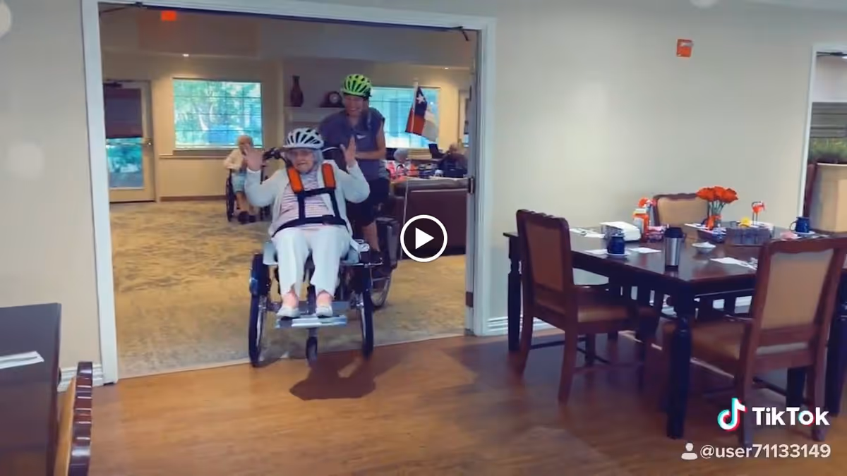 An elderly woman wearing a helmet and safety harness is riding a specialized tricycle indoors, accompanied by a younger person also wearing a helmet. They are moving from a carpeted room into a dining area with a wooden floor and a table set with chairs, cups, and flowers. The setting appears to be a senior living facility.