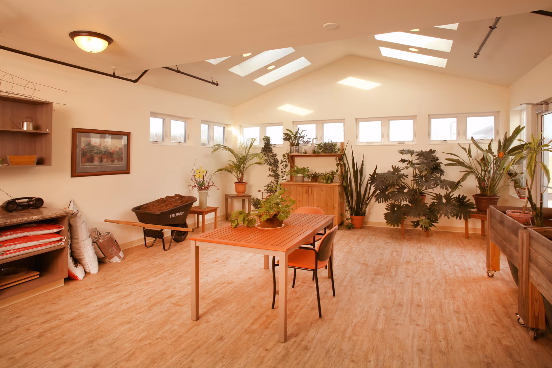A bright indoor gardening room with wooden flooring and white walls. The room features a wooden table with two chairs, several potted plants on shelves and stands, a wheelbarrow filled with soil, bags of gardening supplies, and a wooden cabinet. Skylights and small windows near the ceiling allow natural light to fill the space.