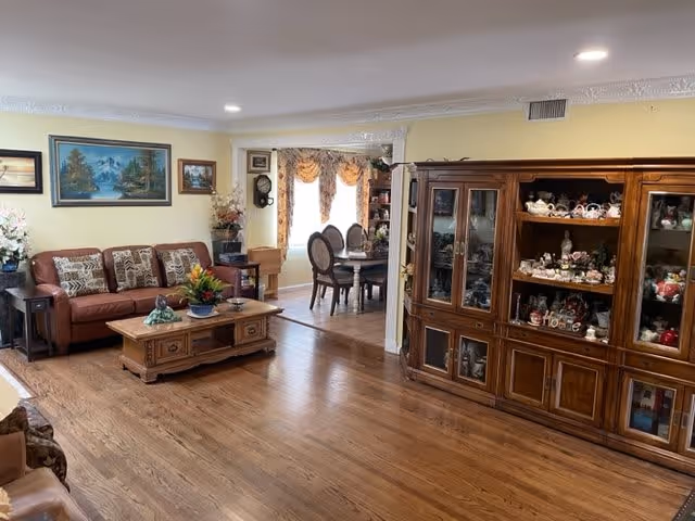 A cozy living room with wooden flooring, a brown leather sofa adorned with patterned cushions, a wooden coffee table with a flower arrangement, and a large wooden display cabinet filled with decorative items. In the background, a dining area with a round table and chairs is visible near windows with ornate curtains.