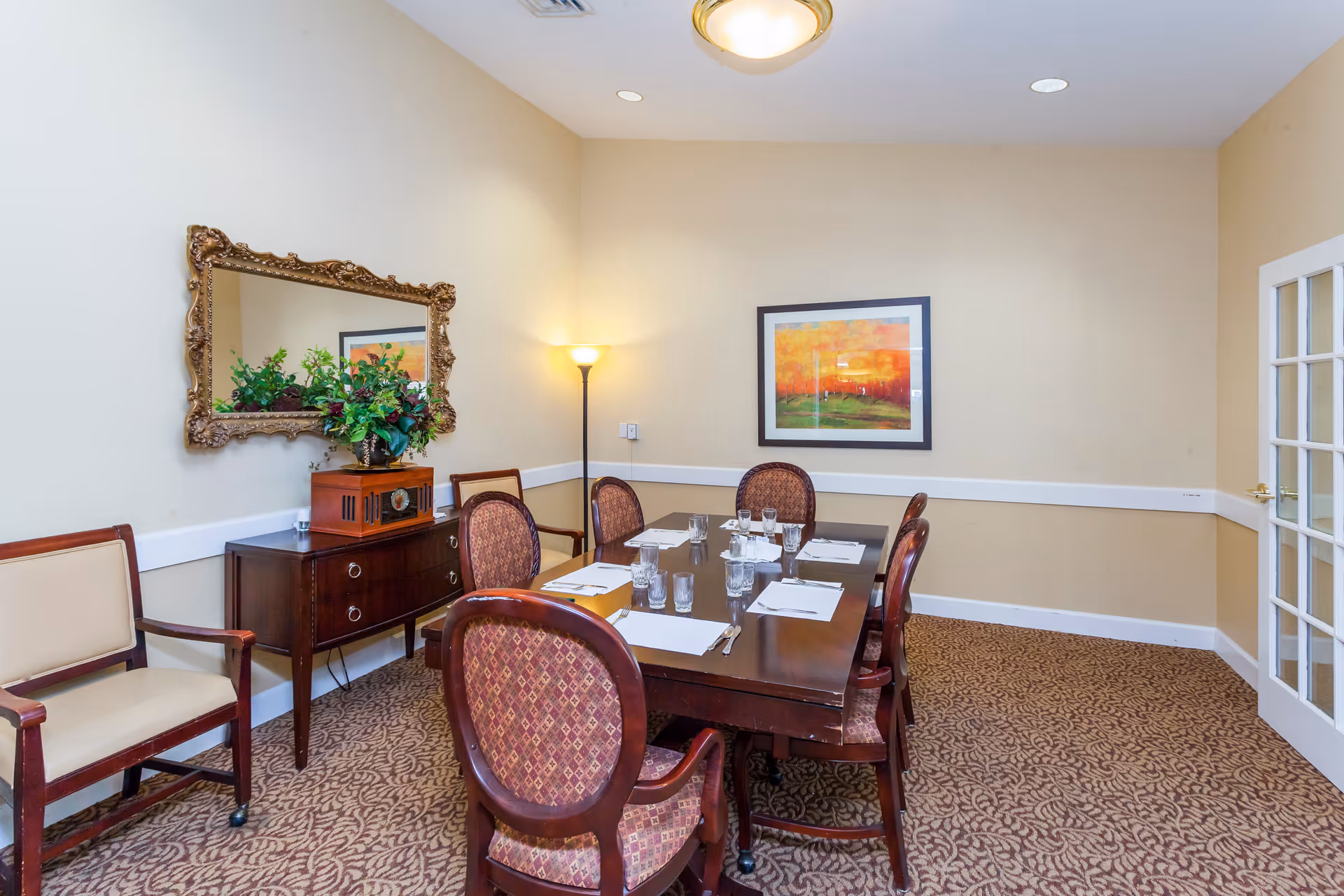 A dining room with a rectangular wooden table set with placemats, glasses, and silverware. Six upholstered chairs surround the table. There is a sideboard with a decorative mirror above it and a floral arrangement on top. A floor lamp is in the corner, and a framed colorful painting hangs on the wall. The room has beige walls and patterned carpet flooring.