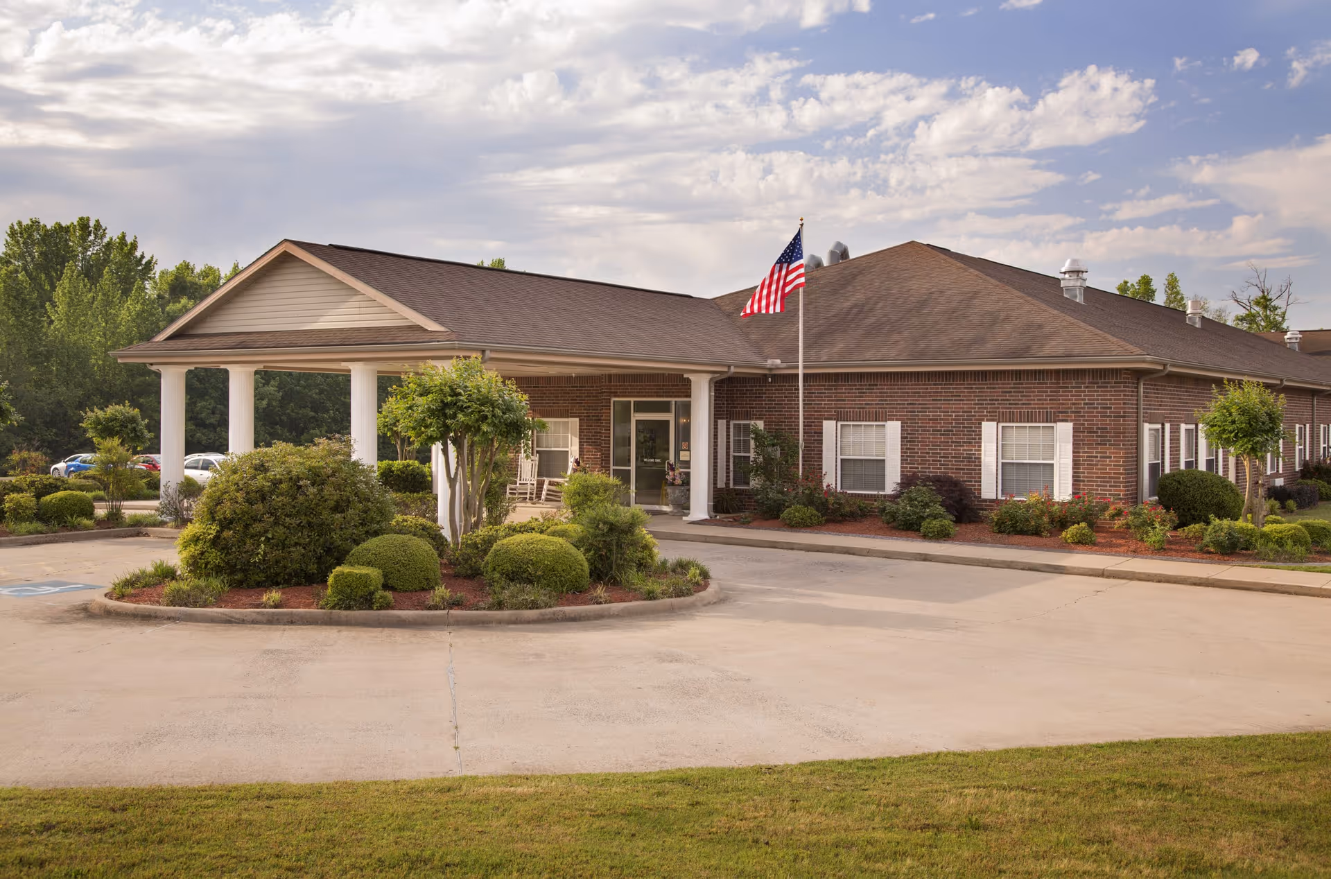 Front exterior of a single-story brick nursing and rehab building with a covered entrance, circular driveway, landscaped shrubs, and an American flag.