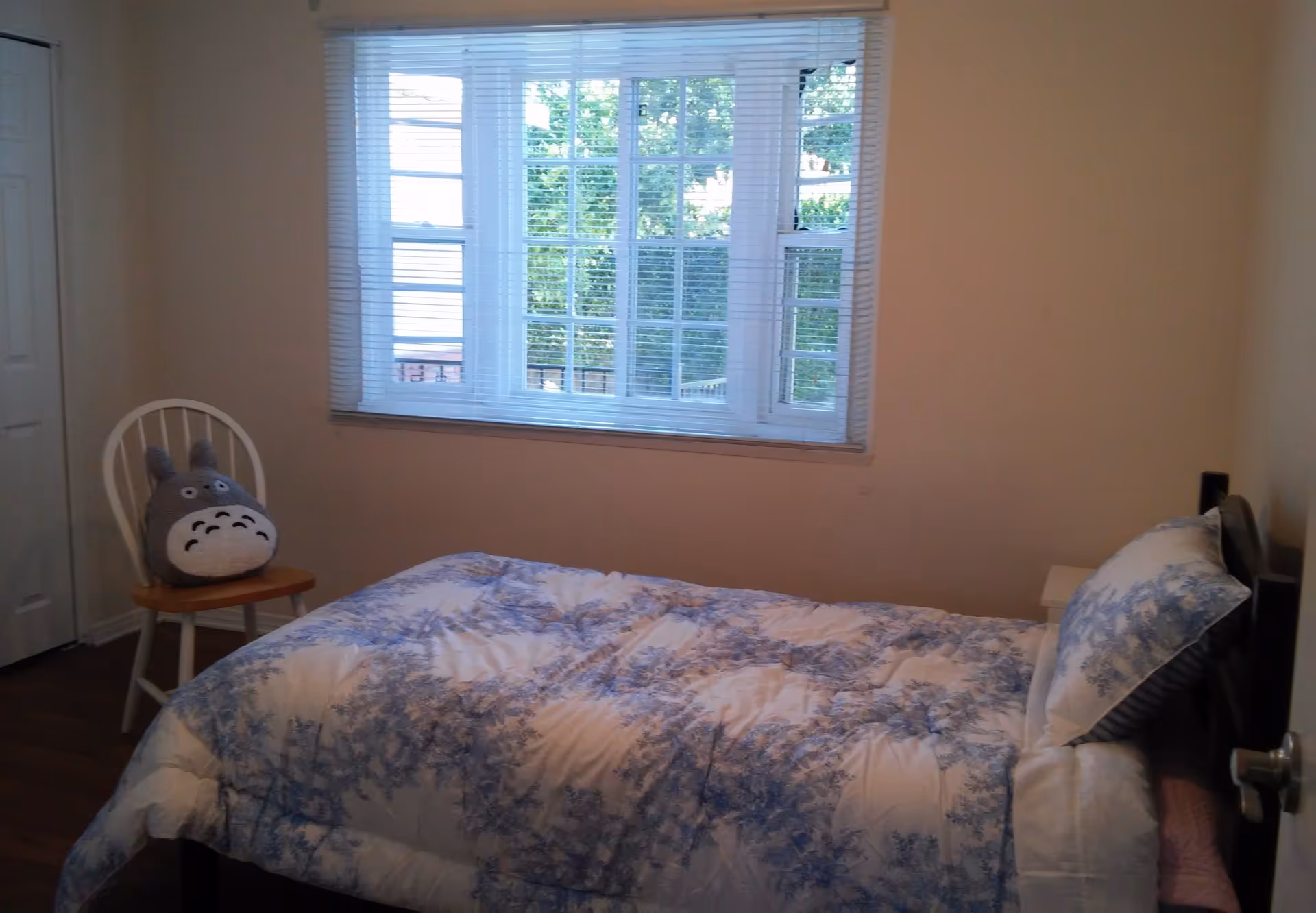 A small bedroom with a single bed covered in a white and blue patterned comforter and matching pillow. There is a white wooden chair with a gray Totoro plush toy on it near a closed closet door. A large window with white blinds lets in natural light, and the walls are painted beige.