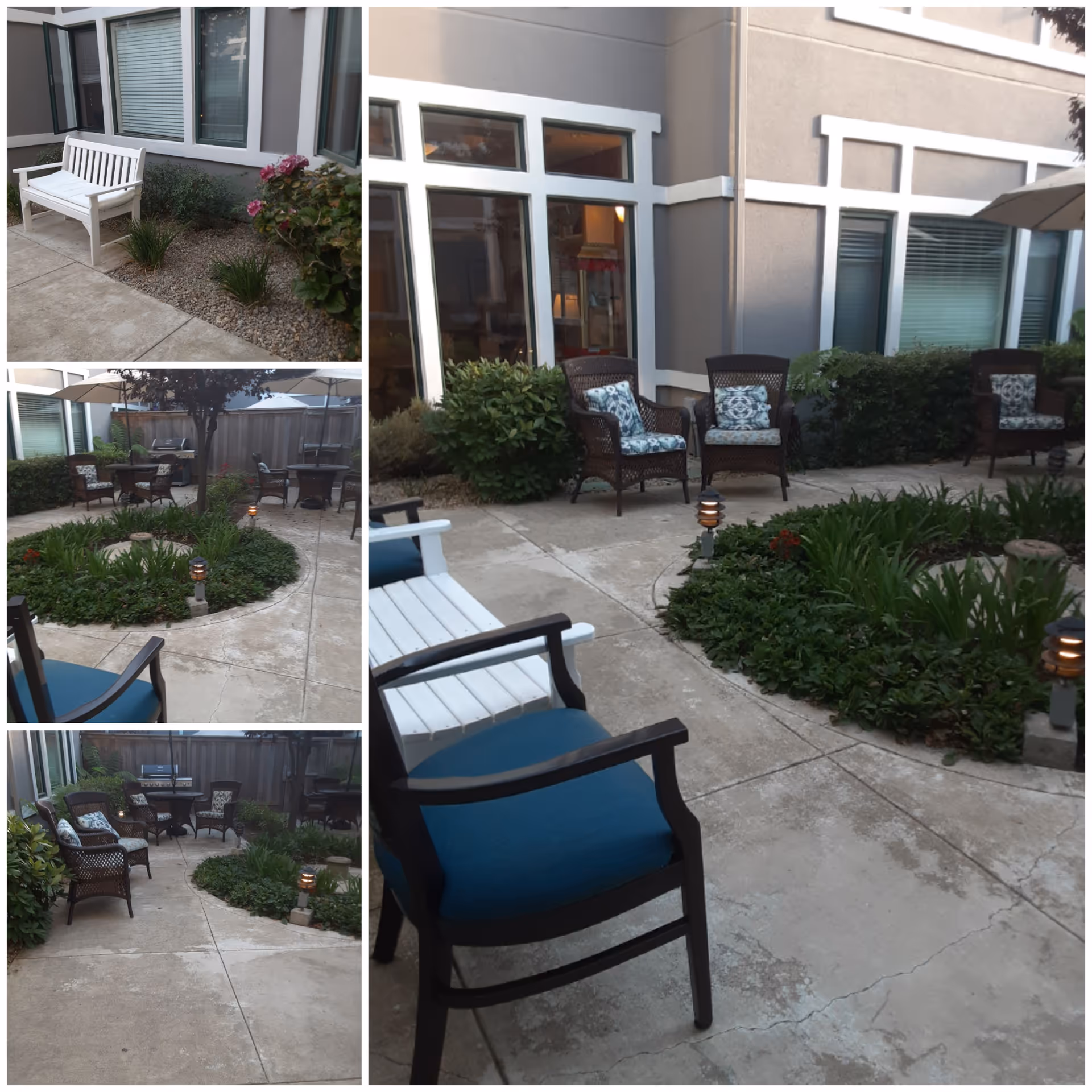 Outdoor patio area with various seating options including white benches and cushioned wicker chairs arranged around a circular garden bed with plants. The patio is surrounded by windows and beige walls of the building, with some small garden lights along the walkway.