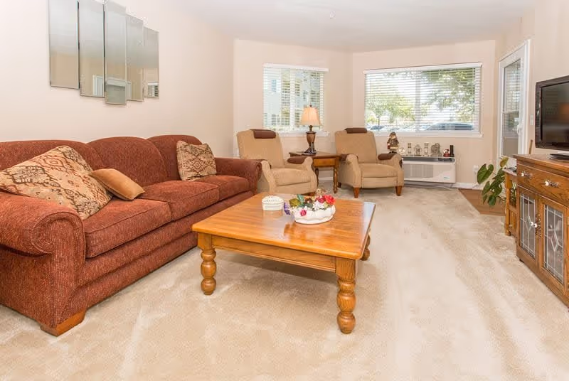 A cozy living room with a brown upholstered sofa adorned with patterned cushions, two beige armchairs, a wooden coffee table with decorative items, a wooden TV stand with a flat-screen television, and windows with white blinds letting in natural light.